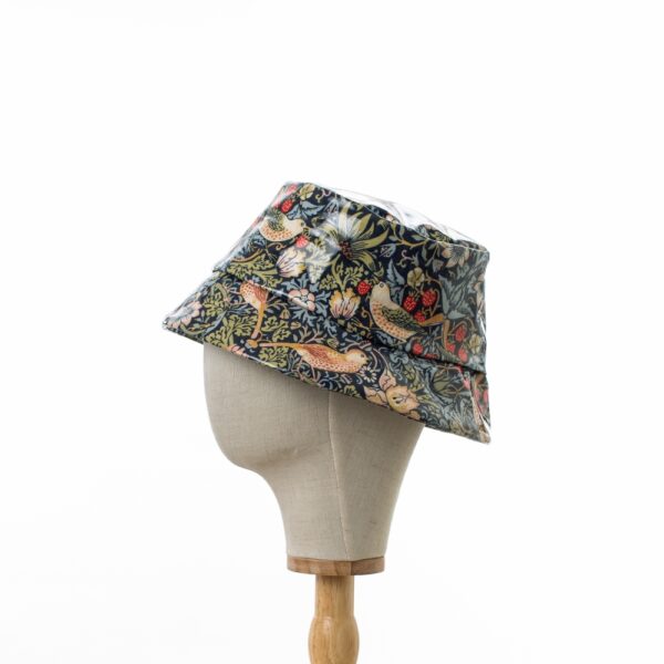 A side on view of a bucket style rain hat in William Morris showerproof fabric on a wooden hat stand. The hat is in the Willilam Morris ‘Strawberry Thief’ design which features thrushes and flowers on a deep navy background