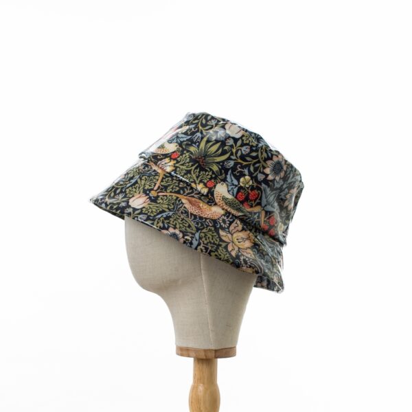 side view of a wider brimmed bucket style rain hat in William Morris showerproof fabric on a wooden hat stand. The hat is made from a glossy pvc fabric in the Willilam Morris ‘Strawberry Thief’ design which features thrushes and flowers on a deep navy background
