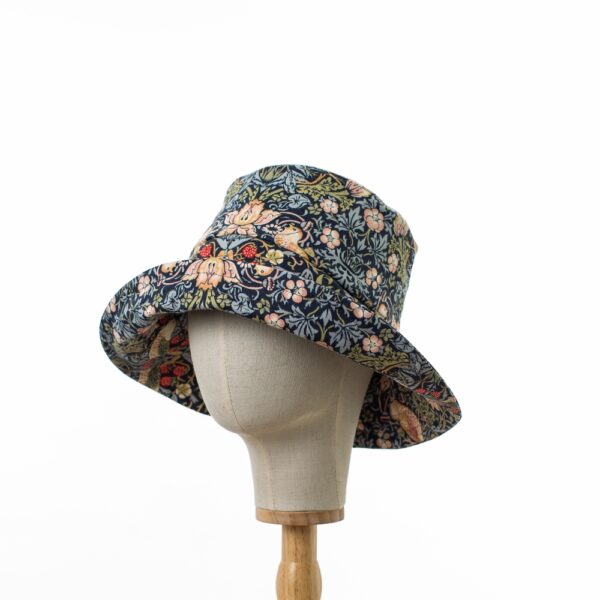 William Morris wide-brimmed hat on a hat stand, featuring an upturned brim. The hat is in the Strawberry Thief pattern with navy fabric, adorned with thrushes, strawberries, and foliage in pink, yellow, and green." The hat stand is turned slightly to the side.