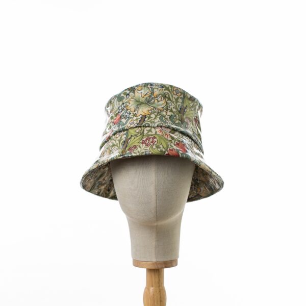 A head on view of a PVC bucket hat featuring William Morris's Golden Lily fabric pattern, with gold and green foliage against a cream-colored background, designed with a glossy finish and displayed on a hat stand.