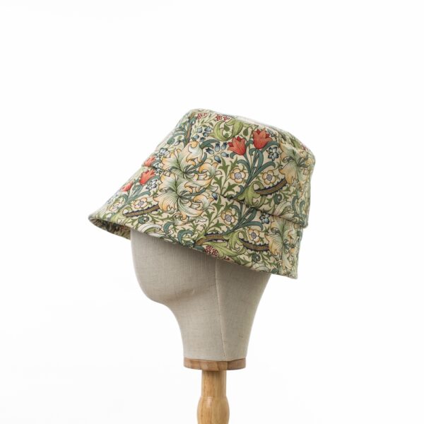 side view of a cotton bucket hat created with William Morris's Golden Lily fabric, featuring a cream background with a pattern of intertwined lilies and foliage in gold and green hues, displayed on a hat stand."