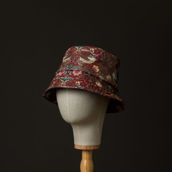 side view of a bucket rain hat in William Morris showerproof fabric. Willilam Morris Red ‘Strawberry Thief’ design which features thrushes and flowers on a deep red background. The image of the hat is on a black background