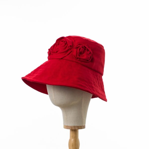 Elegant Bayler and Co wide brimmed Sun Hat with red roses made from 100% red cotton fabric. With the brim upturned and displayed on a canvas and wooden hat stand positioned slightly to the side.