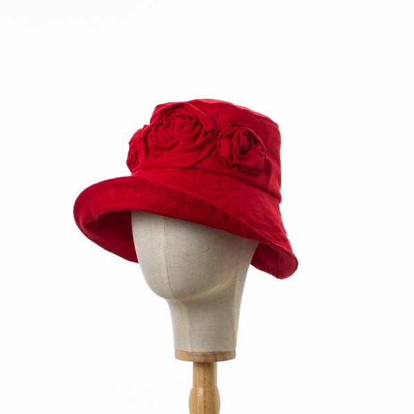 Elegant Bayler and Co wide brimmed Sun Hat with red roses made from 100% red cotton fabric. With the brim upturned and displayed on a canvas and wooden hat stand positioned slightly to the side.