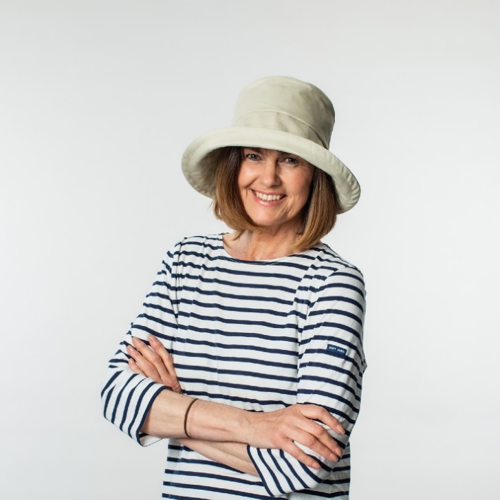 Lucy Bayler from Bayler & Co wearing a cream cotton sunhat with an upturned brim. She is wearing a blue and white striped sailor top with navy denim jeans. She is smiling at the camera with her arms folded.