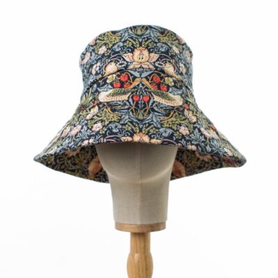 Front view of a William Morris wide brimmed sun hat made from cotton displayed on a stand, showcasing the Strawberry Thief pattern with thrush birds amidst strawberries on a navy backdrop, accented by light blue and green foliage.